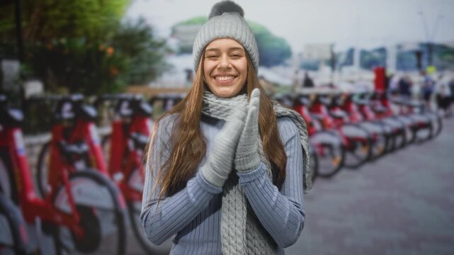 Woman clapping gloved hands beside a row of red rental bikes on a street wearing knit hat and scarf; joyful commute.