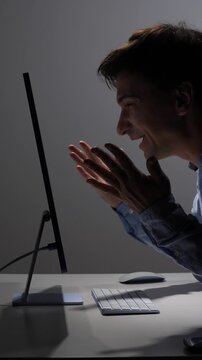 Vertical video man in a shirt celebrating success and achievement while looking at a computer monitor in a dark office, expressing joy and feeling like a winner
