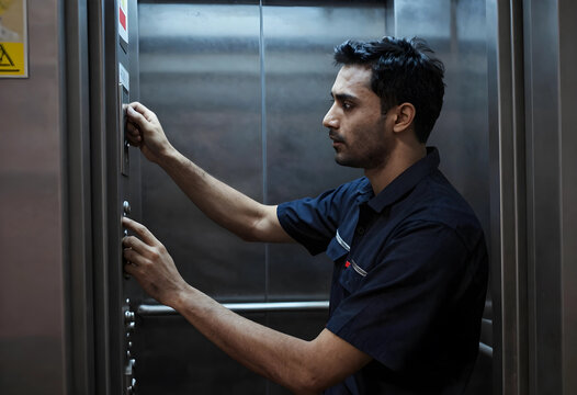 Young male technician in a dark blue uniform performing maintenance on an elevator control panel, the man is focused on the task, using his hands to adjust components and press buttons
