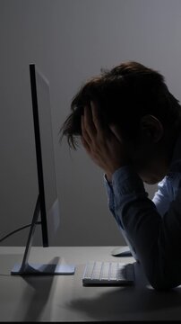Vertical video exhausted person feeling overwhelmed and frustrated, sitting in front of a computer screen, hands covering face in a dark office, symbolizing mental health challenges and fatigue