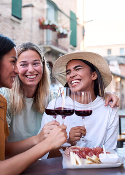 Vertical. Two cheerful multi-ethnic female friends are happily toasting with glasses of red wine while enjoying their vacation together in an outdoor setting in an italian city