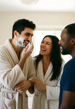 Happy diverse friends in bathrobes laughing together while man applies shaving cream to his face in a bathroom, a man applies shaving cream to his face as a woman stands beside him