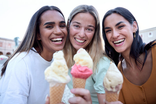 Three cheerful female friends are enjoying a summer day, taking a selfie while laughing and eating ice cream cones in a picturesque town square
