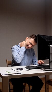 Young man experiencing severe neck pain and discomfort, rubbing his strained muscles while working long hours at a computer in a sedentary office environment