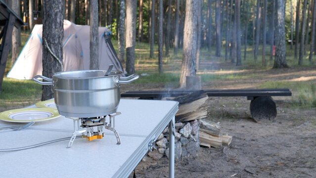 Pot heating on a compact camping stove on a folding table, preparing a meal outdoors at a campsite with a tent and forest in the background