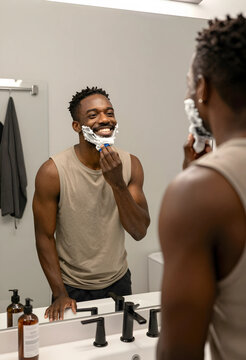 Smiling african american man shaving his face with foam and a razor in a modern bathroom mirror reflection, the mans happy expression reflects a positive start to his day