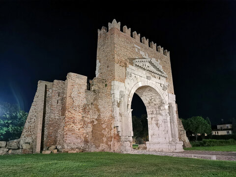 Ancient Roman triumphal Arch of Augustus in Rimini illuminated at night, showcasing intricate stone carvings and historical architecture surrounded by green grass and trees