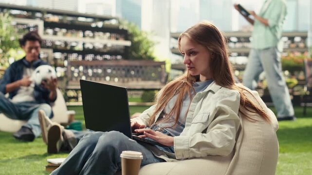 Portrait shows woman focused on writing notes on a laptop while relaxing on a beanbag on a college campus, typing on a computer with stacked books nearby.