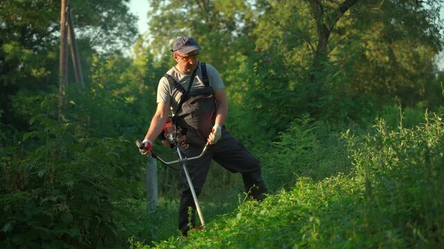 Middle-aged man in gray shirt and black overalls operates a string trimmer to clear tall grass and weeds in a lush green field surrounded by trees and natural vegetation