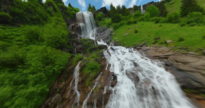FPV aerial view of a waterfall cascading down a lush green mountain slope surrounded by pine trees in Switzerland.