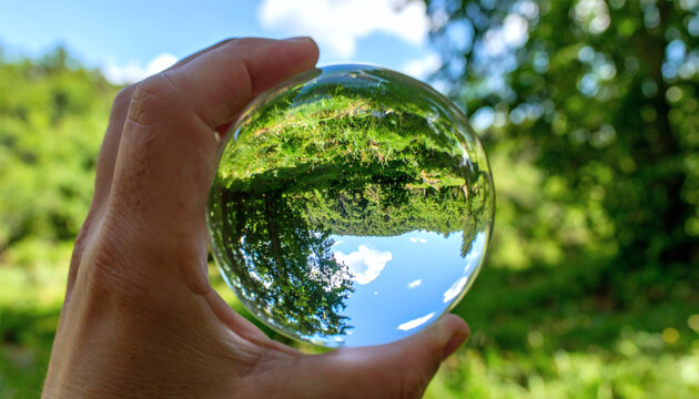 Crystal glass ball held by hand showing a beautiful inverted reflection of green trees and bright blue sky outdoors