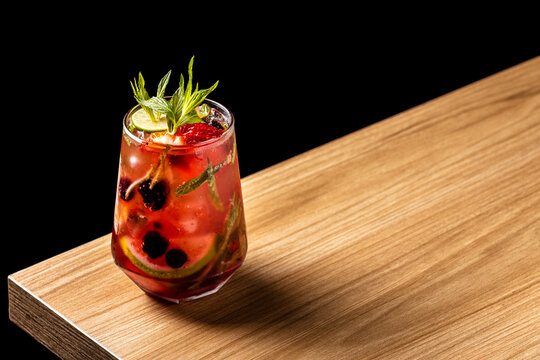 Fruity berry smash cocktail with blackberries, strawberries, mint, and lime on a wooden bar surface with dark background
