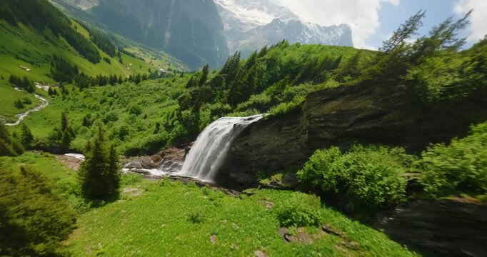 FPV aerial view of a cascading waterfall flowing down rocky cliffs surrounded by lush green alpine meadows and pine trees in Switzerland.