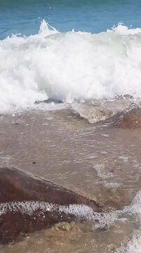 Vertical close-up of foamy coastal surf with clear azure sea waves rolling on empty sand beach and wet boulders at sunny summer day. No people seaside water surface pattern natural background.
