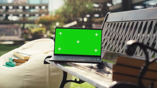 Green screen chroma key on a laptop appears on a wooden bench, offering an isolated editable screen for education and digital learning visuals, with an empty campus landscape and mockup screen.