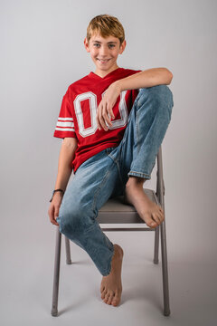 Smiling barefoot boy wearing red American football jersey and jeans sitting with one leg up on a folding chair
