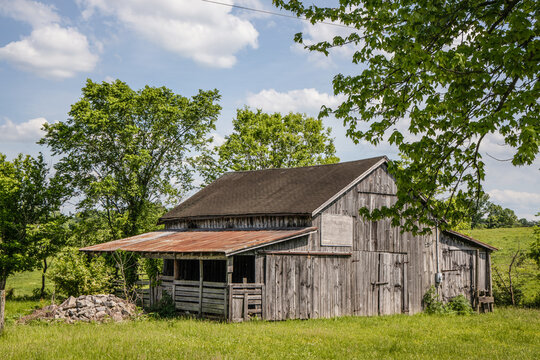 View of a weathered wooden barn with a rusty metal roof, set in a lush green field surrounded by trees under a bright blue sky in Kentucky, United States.
