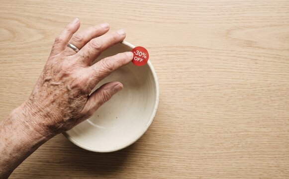 Elderly person hand touching a ceramic bowl with a minus thirty percent off discount sale tag on a wooden table