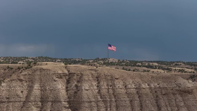 Aerial of American flag over arid badlands landscape in Duchesne Utah under stormy dark sky with rolling hills and deep eroded cliffs
