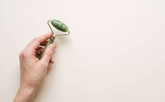 Hand holding a green jade facial roller for skin care and wellness massage on neutral background