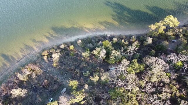 Wooded shoreline borders tranquil blue water near Hawthorn Circle park grounds. Sunlit trees shade small campsites along sandy banks of Lake Ray Roberts State Park, Pilot Point, Texas