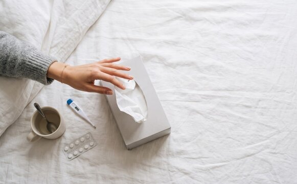 Sick woman in bed reaching for a tissue box with medicine and thermometer on white sheets