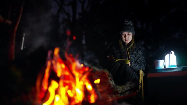Asian woman writing travel journal and drinking hot coffee beside campfire and tent in forest mountain. People relax and enjoy travel outdoor active lifestyle hiking, backpacking and nature healing.