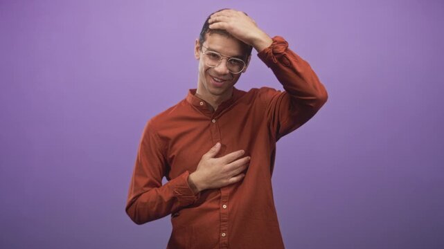 Young hispanic man touches chest with one hand and touches head with the other in studio purple backdrop; shy gratitude.