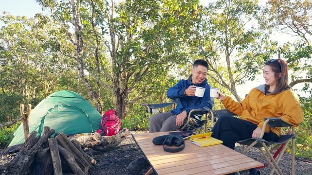 Happy Asian couple drinking hot coffee while travel nature and camping at forest mountain. Man and woman relaxing at campsite, enjoying morning beverage with outdoors active lifestyle nature therapy.
