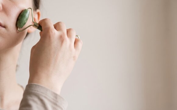 Young woman using a green jade facial roller for skin care and lymphatic massage at home