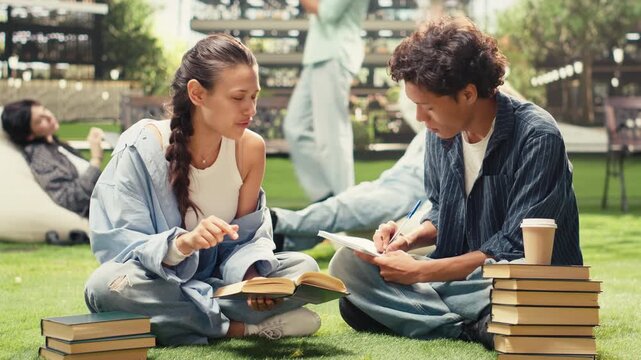 Diverse classmates collaborate with textbooks while seated on a university campus lawn, discussing coursework and taking notes for exam preparation, supporting peer tutoring and group study.