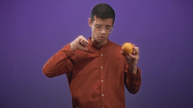 Man holds orange fruit in right hand and shows left thumbs down with puzzled look in studio; skepticism tasting critique.