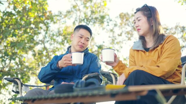 Happy Asian couple drinking hot coffee while travel nature and camping at forest mountain. Man and woman relaxing at campsite, enjoying morning beverage with outdoors active lifestyle nature therapy.