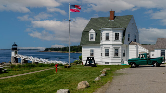 Marshall Point Lighthouse with green roof sits on the coast in Maine, accompanied by an American flag. The scene includes a classic green truck parked on the driveway.