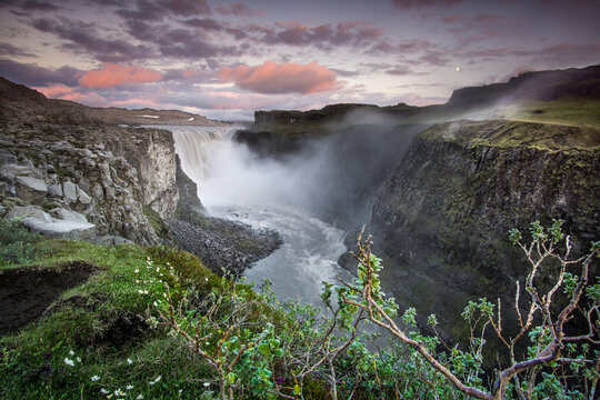 View of Dettifoss waterfall cascading into a deep rocky canyon with lush green vegetation in the foreground under a dramatic sunset sky in Nordurthing, Iceland.
