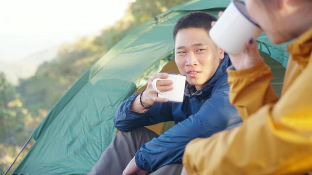 Happy Asian couple drinking hot coffee while travel nature and camping at forest mountain. Man and woman relaxing at campsite, enjoying morning beverage with outdoors active lifestyle nature therapy.