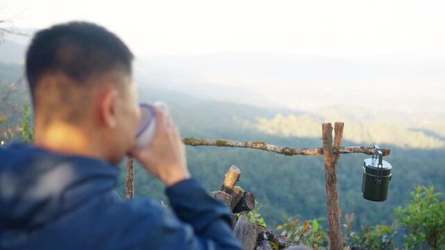 Asian man drinking hot coffee while solo travel nature and camping at forest mountain. Male backpacker relaxing at campsite, social detox, enjoy nature therapy morning beverage with outdoor lifestyle