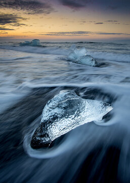 View of ice chunks on the black sand beach of Diamond Beach at Jokulsarlon Glacier Lagoon with waves washing over the ice under a sunset sky in Hornafjordur, Iceland.