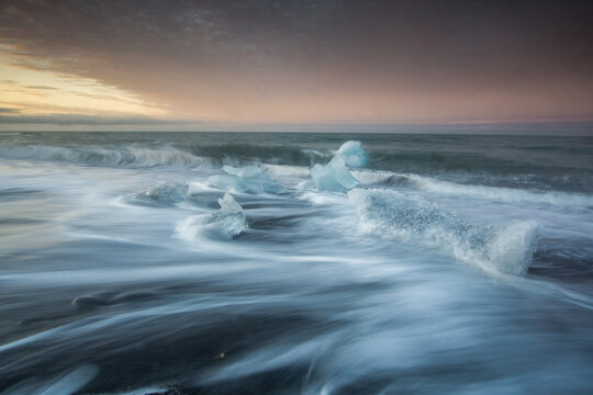View of ice chunks from Jokulsarlon Glacier Lagoon washing up on the black sand beach at sunset with long exposure waves, Sveitarfelagid Hornafjordur, Iceland.