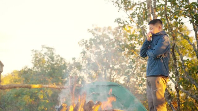 Asian man drinking hot coffee while solo travel nature and camping at forest mountain. Male backpacker relaxing at campsite, social detox, enjoy nature therapy morning beverage with outdoor lifestyle