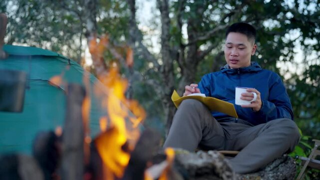 Asian man writing travel journal and drinking hot coffee beside campfire and tent in forest mountain. People relax and enjoy travel outdoor active lifestyle hiking, backpacking and nature healing.