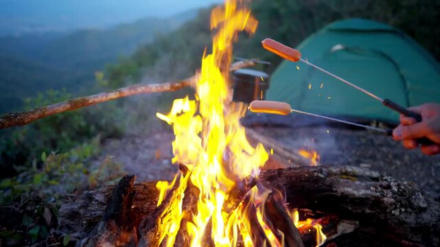 Happy Asian family couple travel nature camping on mountain peak sitting near campfire roasting sausage and drinking beer together at night. People enjoy outdoor active lifestyle and nature therapy