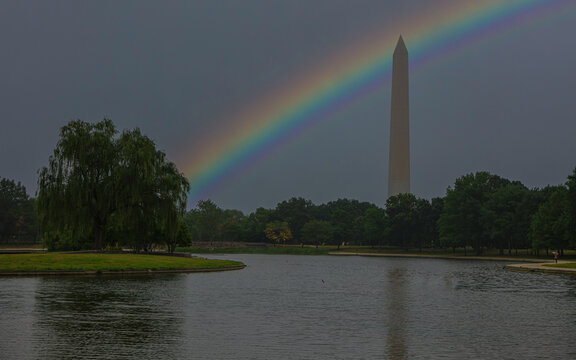A vibrant rainbow arches over the Washington Monument, creating a stunning contrast against the gray sky. The reflection of trees by the Constitution Gardens Pond adds to the peaceful atmosphere.