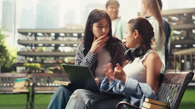 Team of university students sits on a bench with an open laptop and stacked books, smiling as they discuss project ideas in a modern campus courtyard, with classmates studying together.
