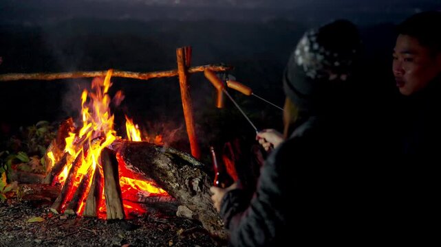 Happy Asian family couple travel nature camping on mountain peak sitting near campfire roasting sausage and drinking beer together at night. People enjoy outdoor active lifestyle and nature therapy