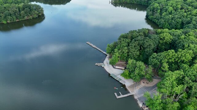 Aerial drone footage flying over a calm lake towards a boat launch area with wooden docks. The shoreline is bordered by a dense green summer forest in a tranquil park setting in Burke, Virginia.