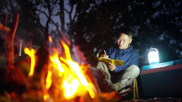 Asian man writing travel journal and drinking beer beside campfire and tent in forest mountain. People relax and enjoy travel outdoor active lifestyle hiking, backpacking and nature healing.