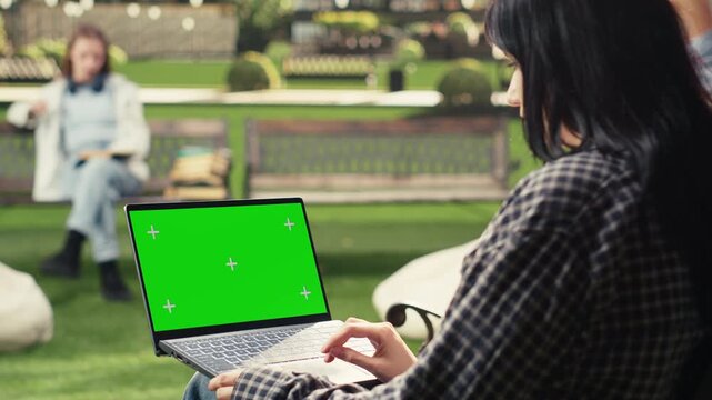 Green screen phone mockup is held close to the camera in an outdoor campus setting, with a blurred student in the background and copy space for mobile screen replacement and digital education content.