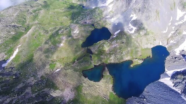 Aerial drone view of alpine tarn lake with rocky shore, green meadow, and mountain peaks.