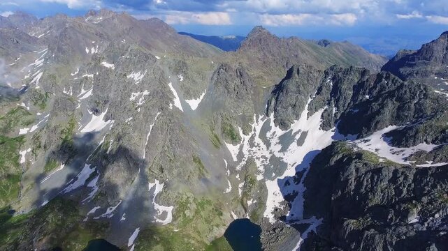 Aerial drone view of alpine tarn lake with rocky shore, green meadow, and mountain peaks.